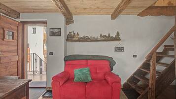 A red armchair with a green pillow in a room with wooden beams, a staircase, and a wall-mounted shelf with a landscape picture.