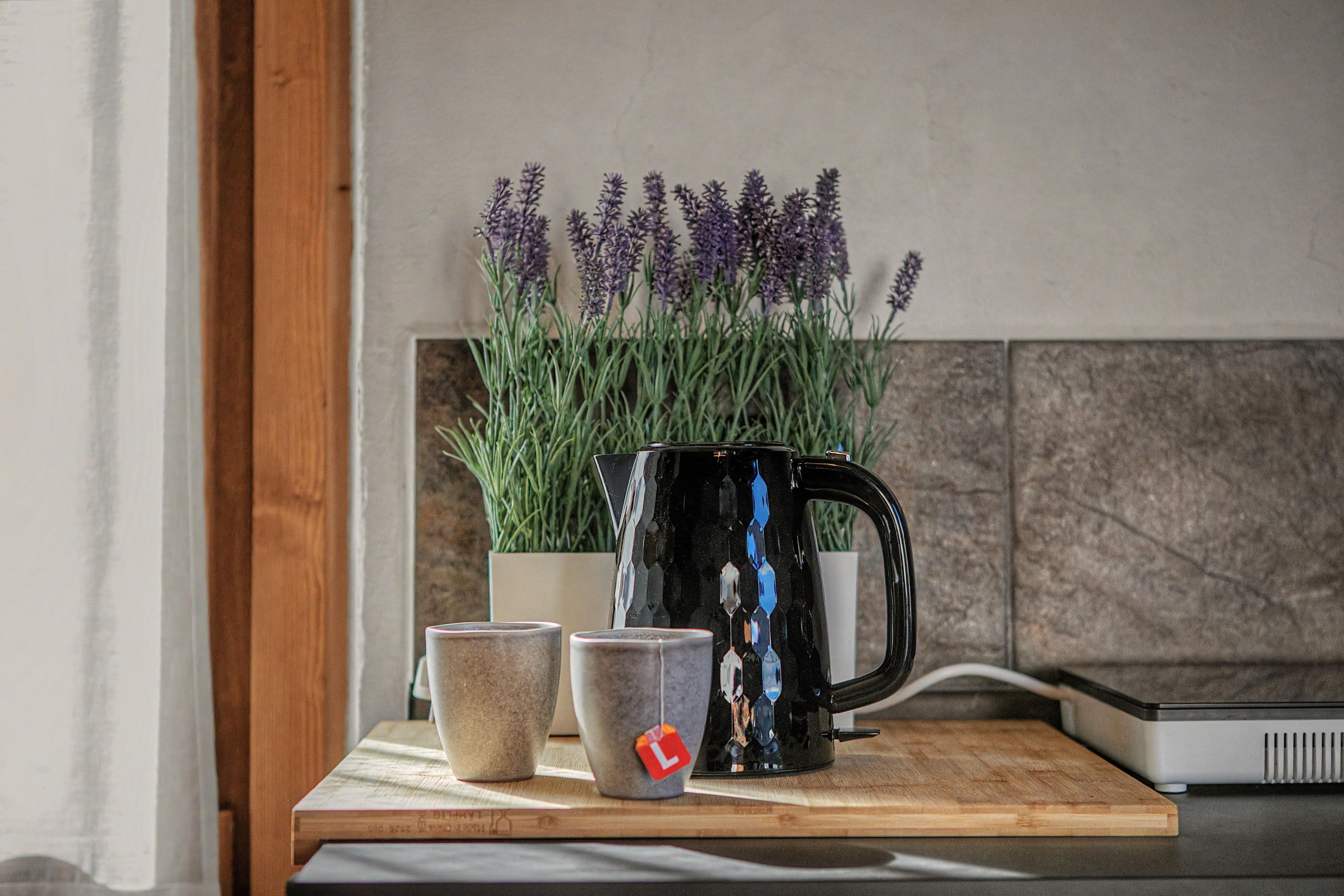 Eine Küchenarbeitsplatte mit einer Kaffeemaschine, zwei Tassen und einem Blumentopf mit violetten Blüten.