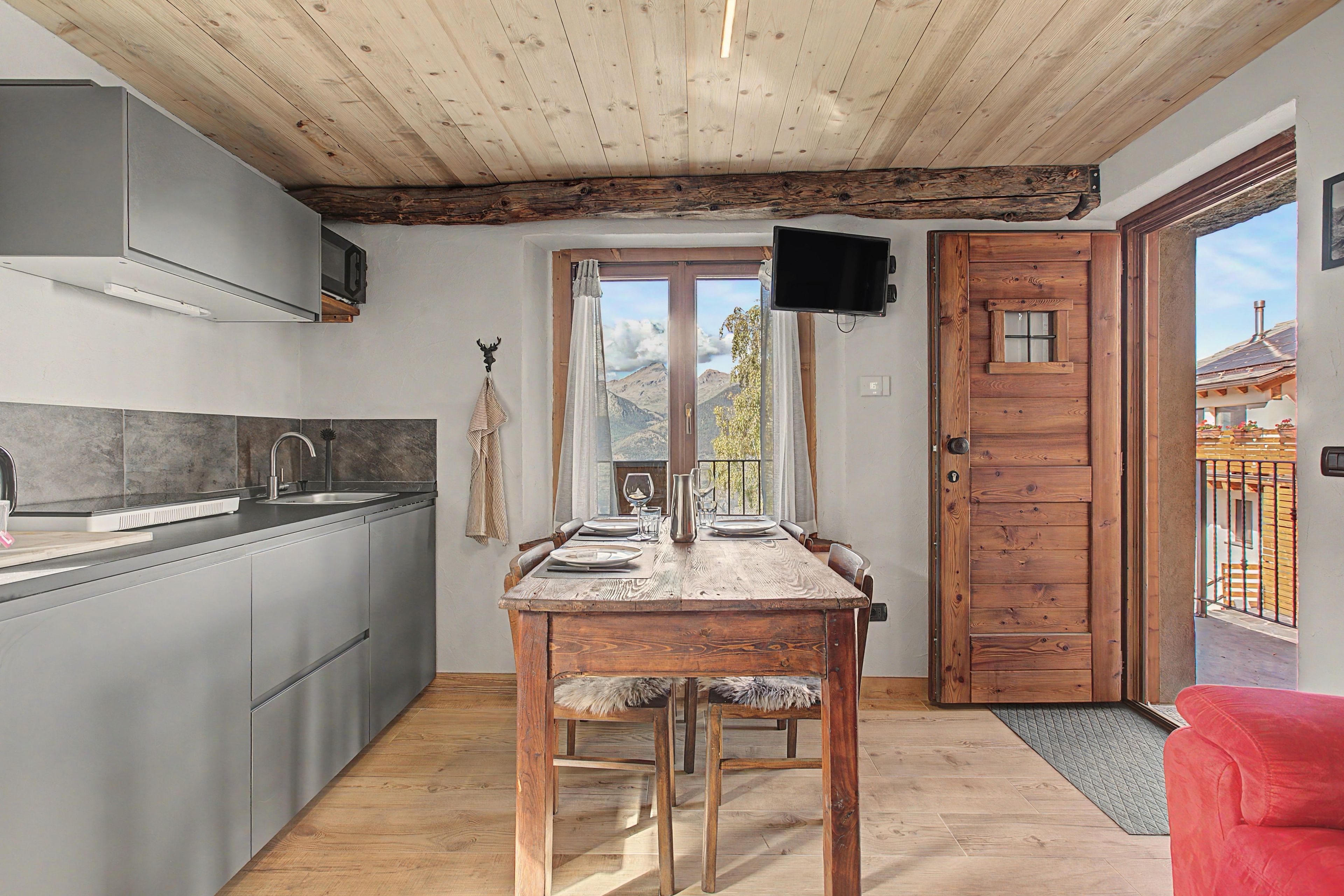 Una cocina con una mesa y sillas de madera, un televisor en la pared y vistas a las montañas por la puerta.