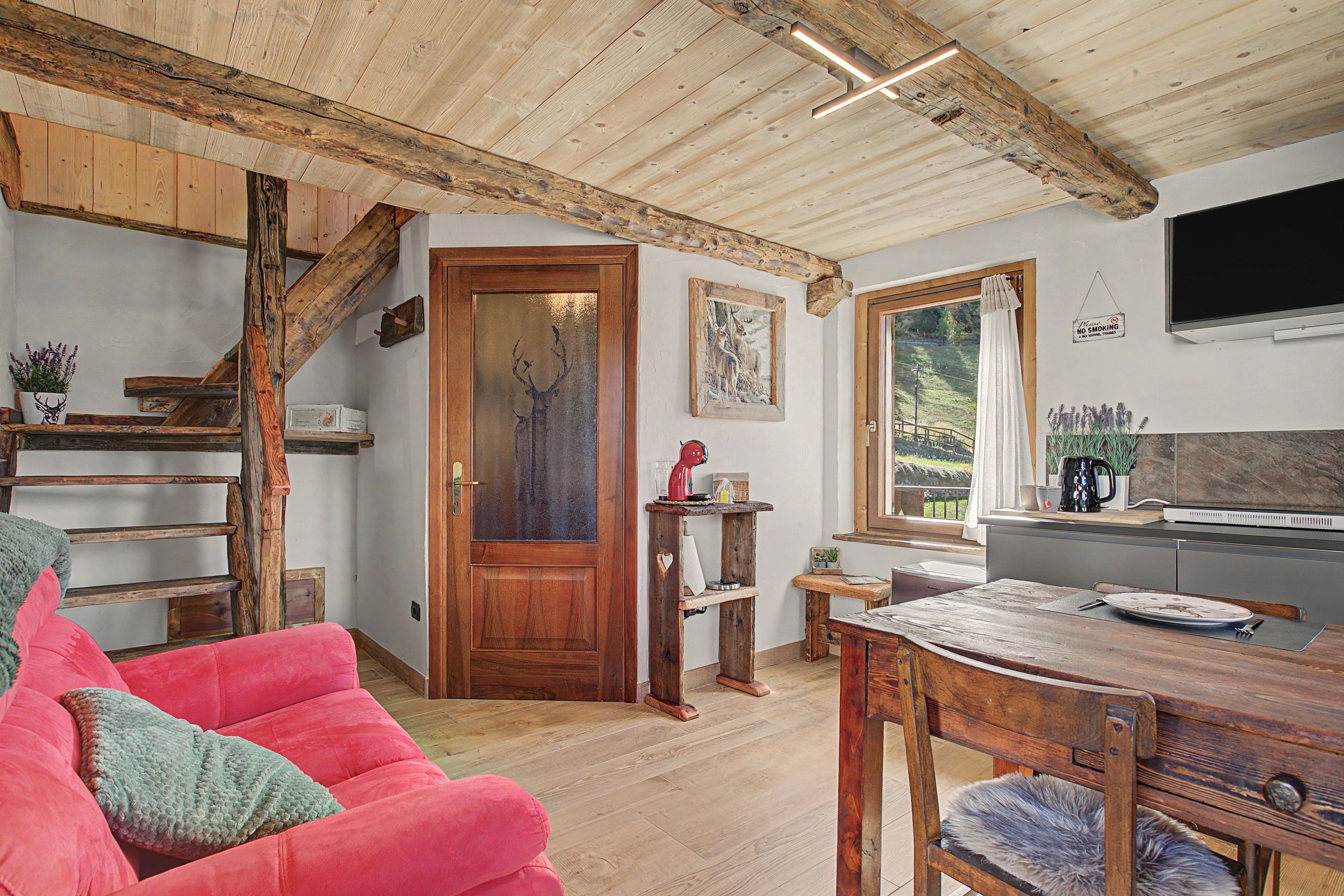 Een rustiek interieur met een houten trap, een rode bank, een eettafel en een keukenhoek.