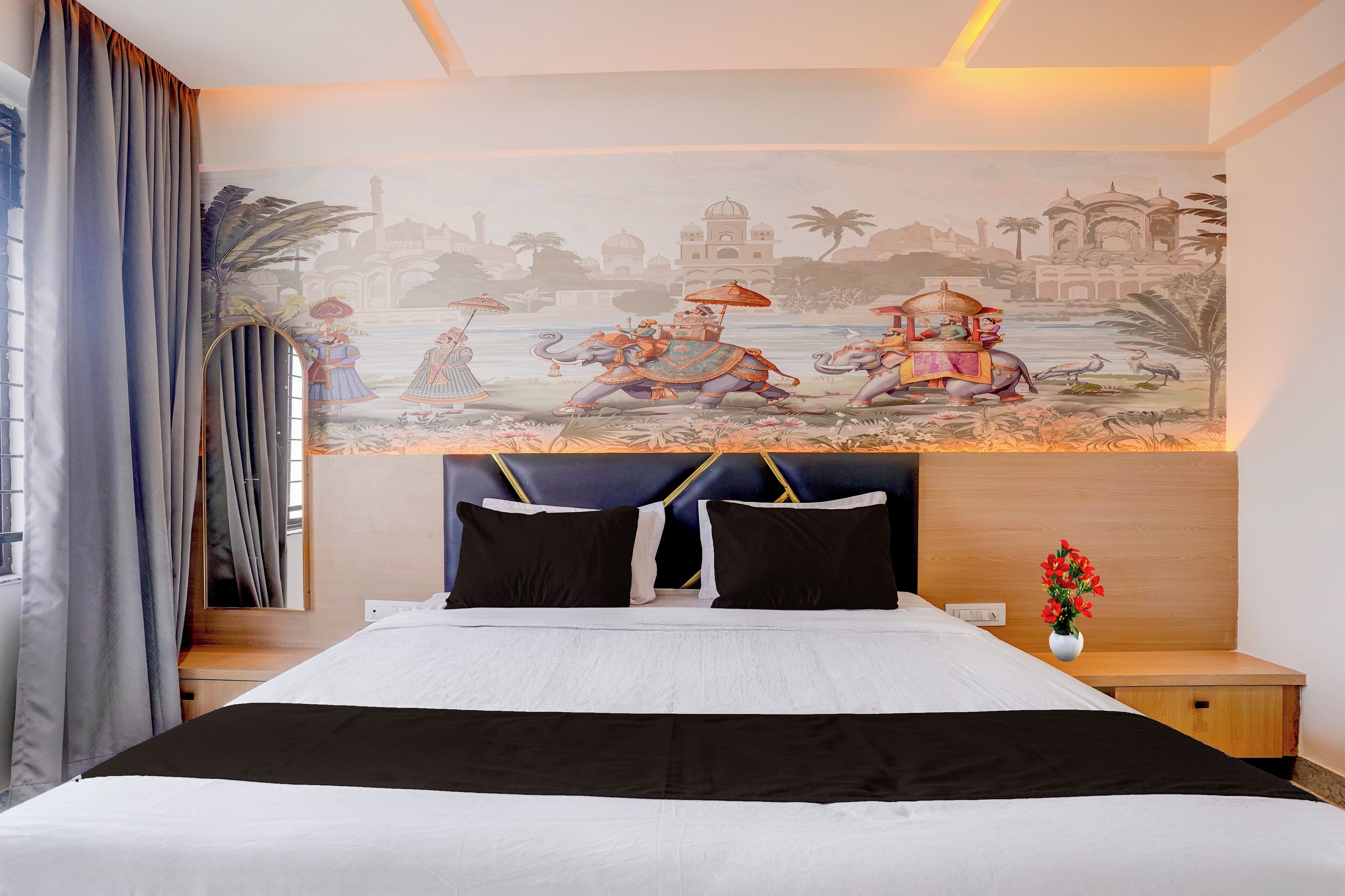 Een hotelkamer met een grote muurschildering van een traditioneel tafereel, een bed met zwart-wit beddengoed en een nachtkastje met een bloevas.