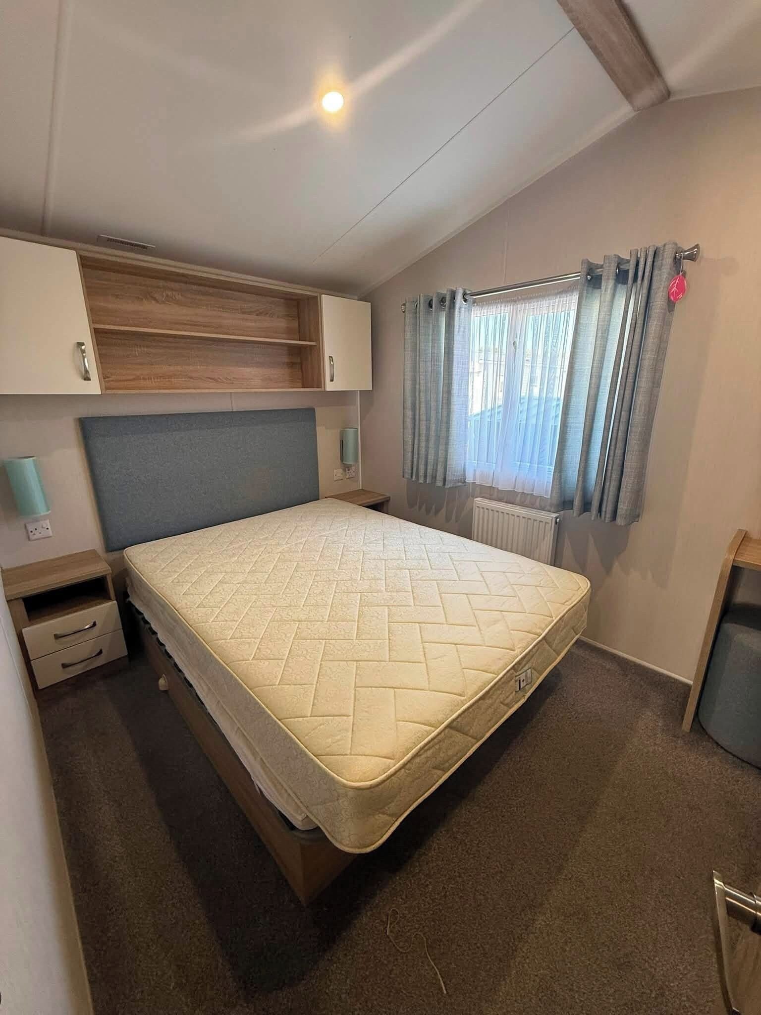A bedroom with a bed, a wooden headboard, a nightstand, a wall-mounted lamp, a window with curtains, and a radiator.