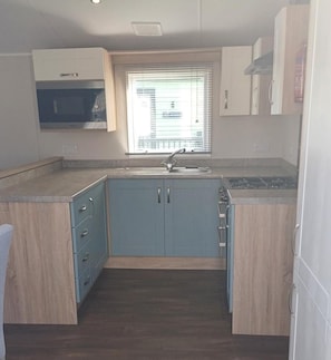 A compact kitchen with a built-in sink, a window with blinds, and light-colored cabinetry. - Home by the Sea - Adventure Village Access (Great Yarmouth)