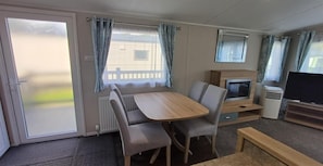 A compact living space with a dining table, chairs, a flat-screen TV, and a fireplace. - Home by the Sea - Adventure Village Access (Great Yarmouth)