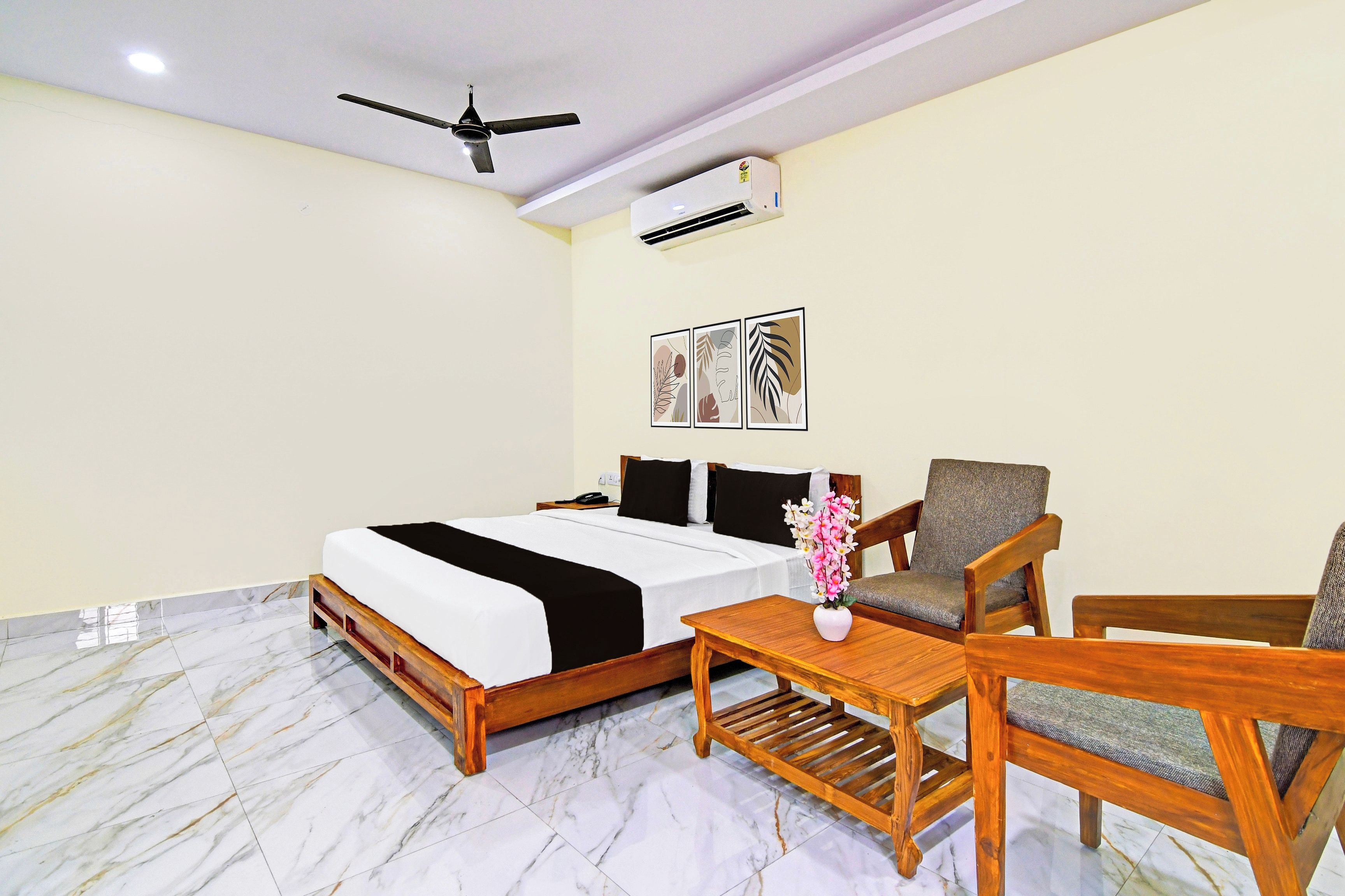 A hotel room with a bed, a desk, two chairs, a ceiling fan, an air conditioner, and wall art.