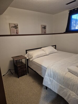 Una cama individual con estructura de madera, una mesita de noche pequeña y un estante en la pared con dos fotografías enmarcadas.