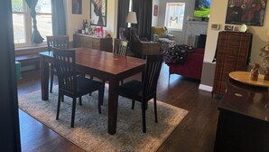 Spiseplads med et træbord og stole, et køkken i baggrunden og en stue med en rød sofa.