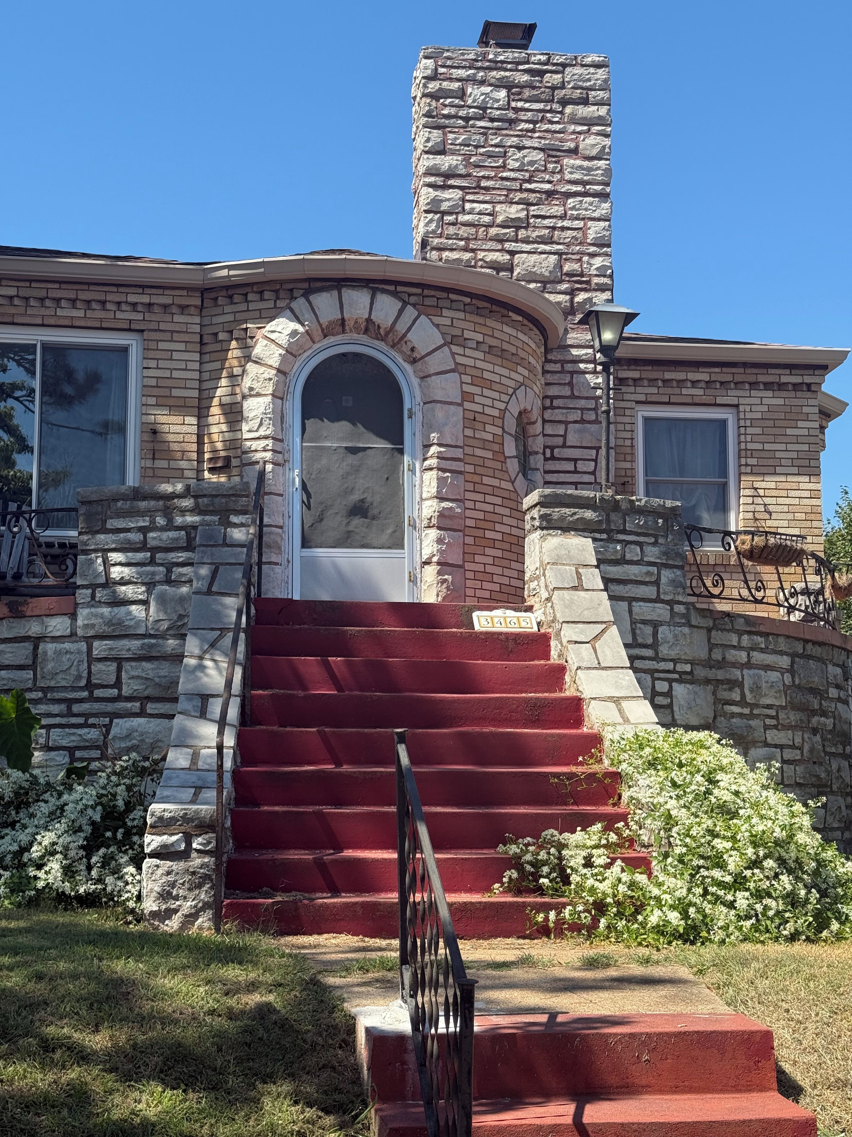 Une maison en pierre avec un escalier rouge menant à une porte blanche, le tout entouré de verdure.