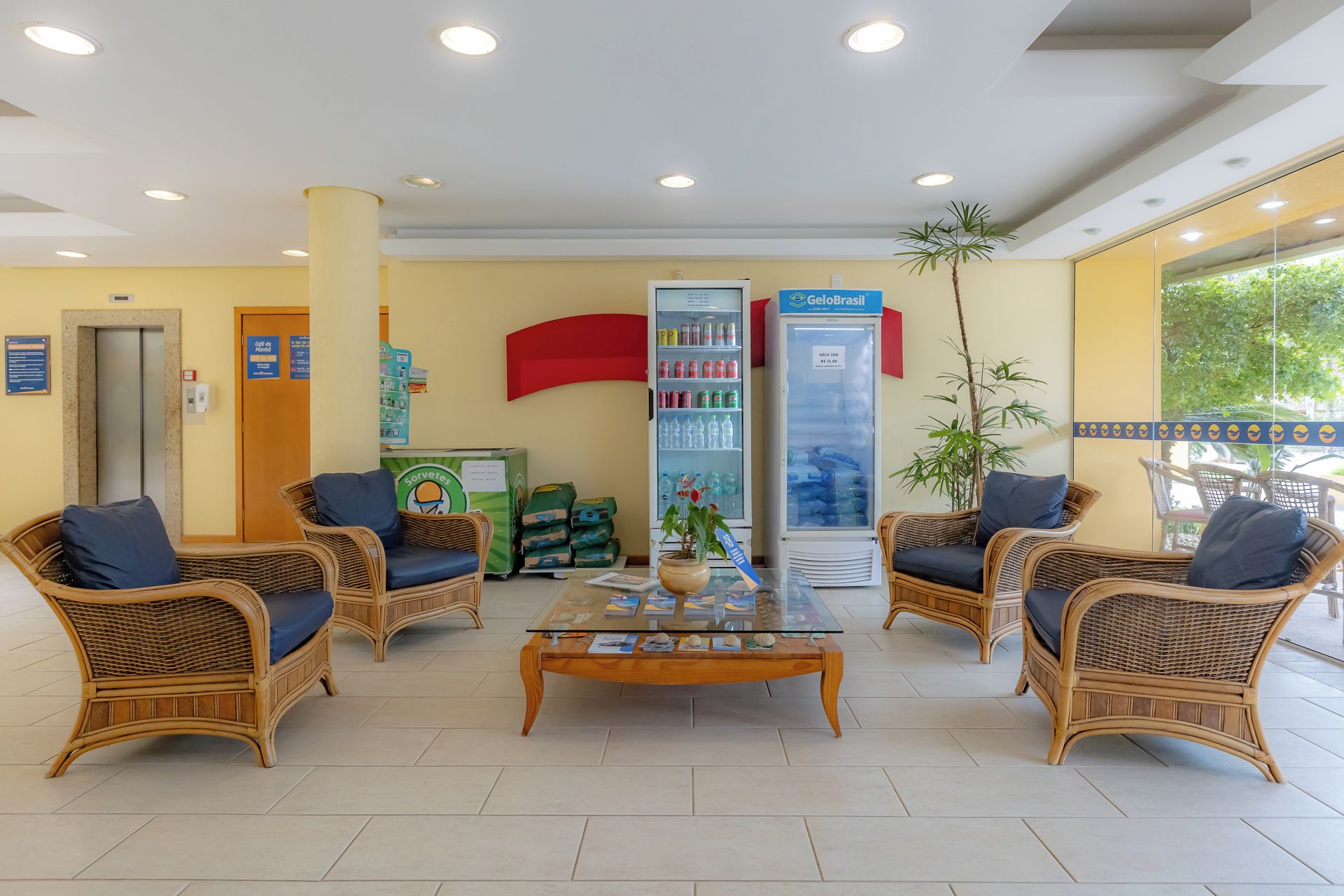 A reception area with wicker furniture, a glass coffee table, a refrigerator, and a plant.
