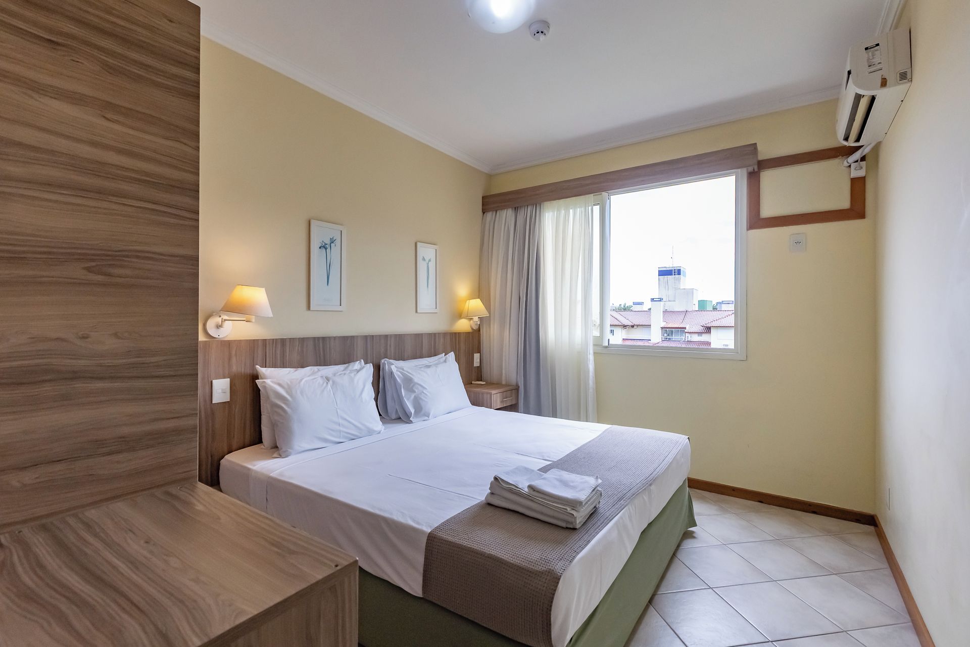 A hotel room with a bed, bedside lamps, a window with curtains, and a view of buildings outside.