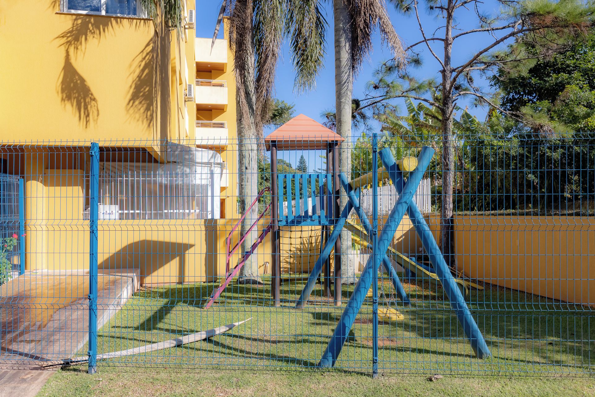 A fenced outdoor area with a playground structure, including swings and a climbing frame, adjacent to a yellow building with multiple balconies.