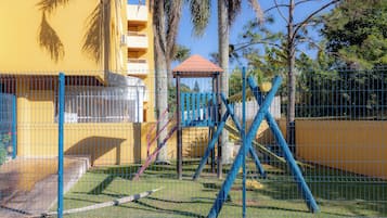 Un espacio exterior cercado con una estructura de juegos, que incluye columpios y un gimnasio de escalada, junto a un edificio amarillo con varios balcones.