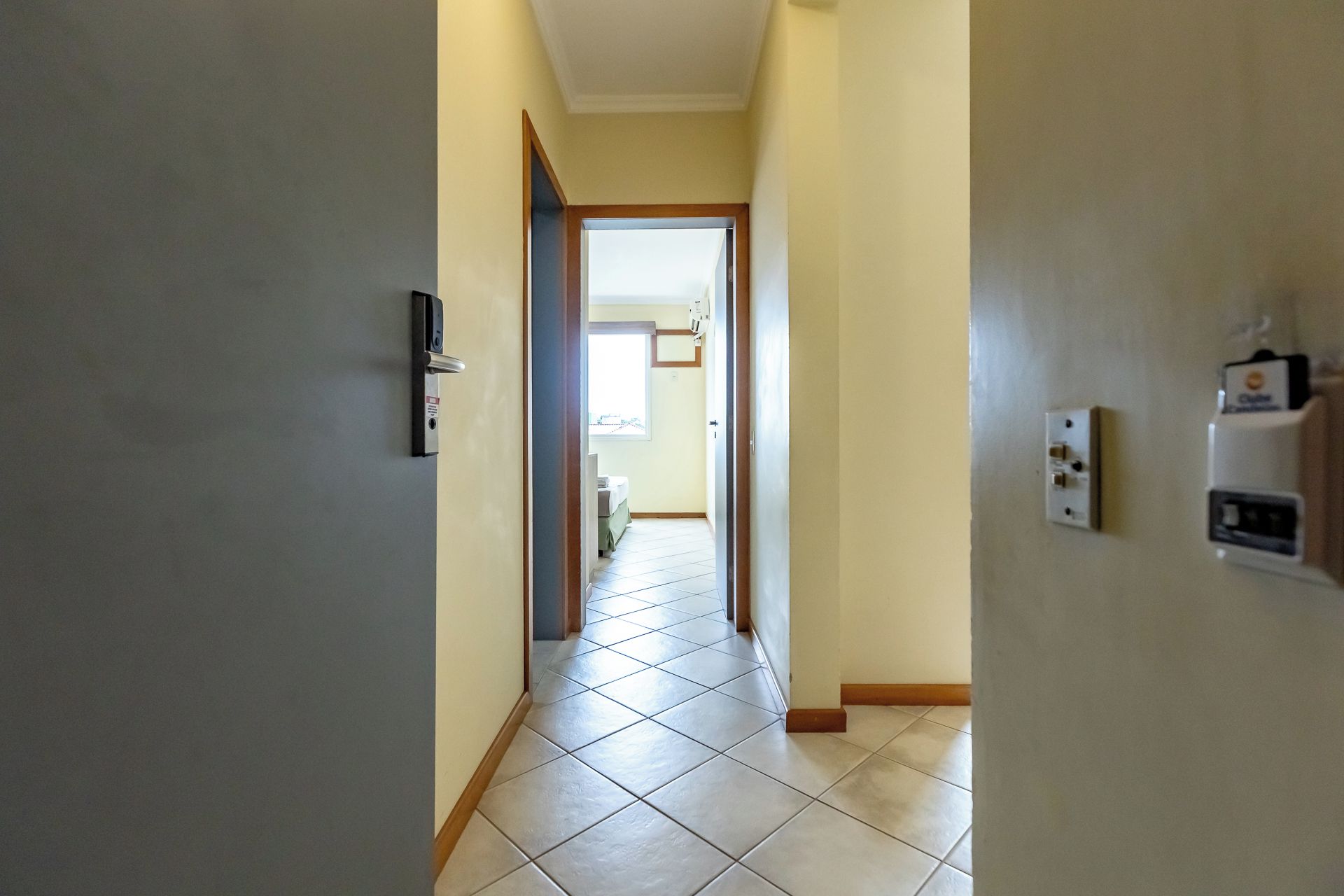 Um corredor com uma porta à esquerda, um interruptor de luz à direita e vista para outro cômodo.