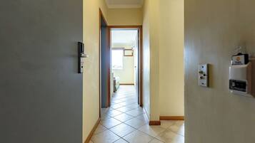 Um corredor com uma porta à esquerda, um interruptor de luz à direita e vista para outro cômodo.