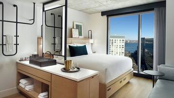 Una habitación de hotel moderna con una cama grande, una mesita de noche con un lavamanos, un espejo y vistas de la ciudad a través de un ventanal.