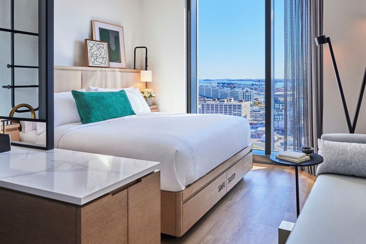 Une chambre d’hôtel moderne équipée d’un lit, d’une table de chevet, d’une lampe et offrant une vue sur le paysage urbain.