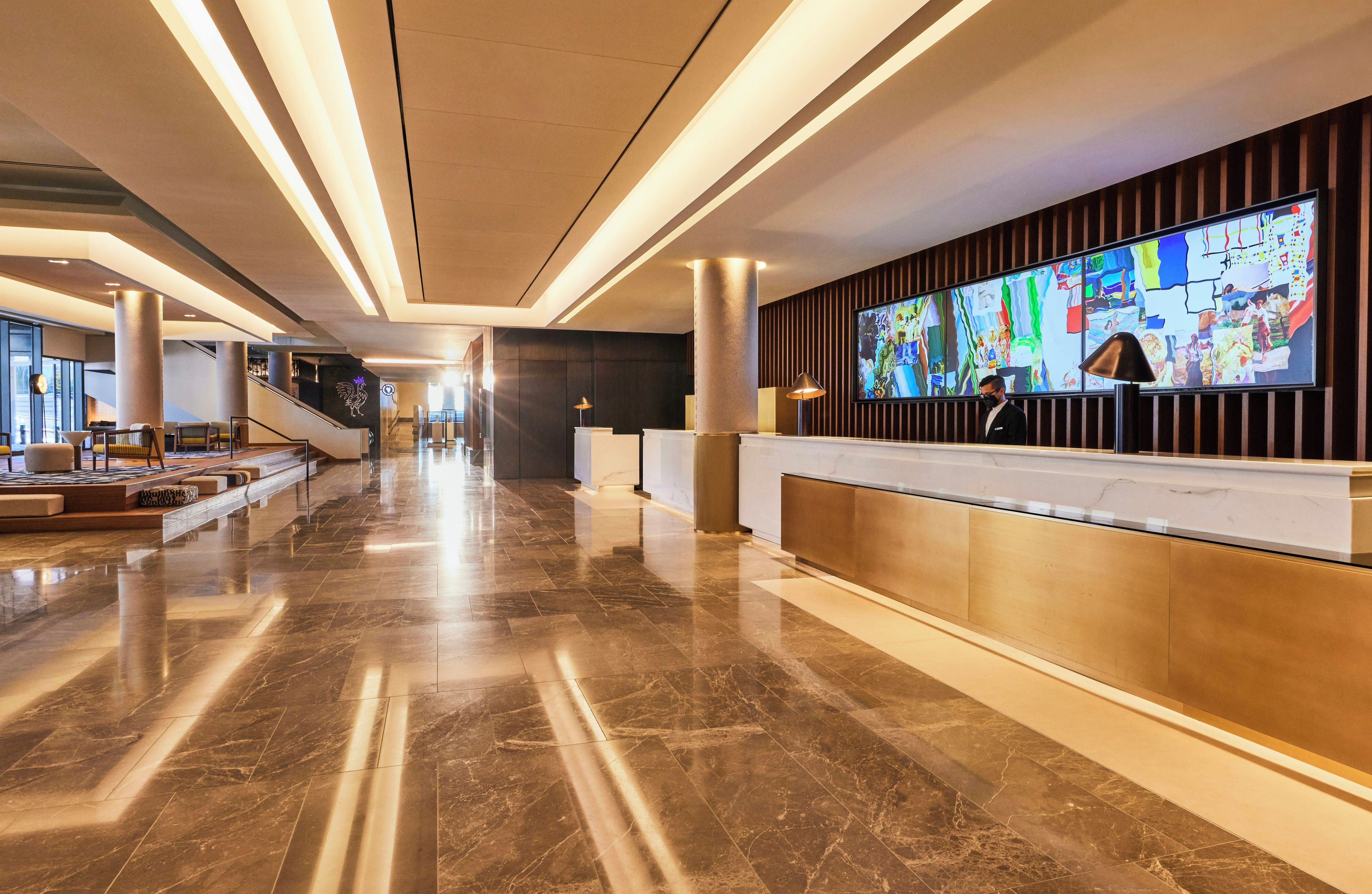 A modern hotel lobby with a long reception desk, marble flooring, and a large digital display.