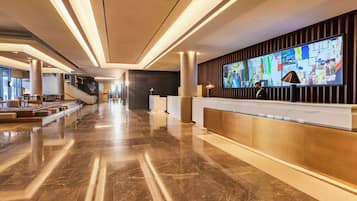 A modern hotel lobby with a long reception desk, marble flooring, and a large digital display.
