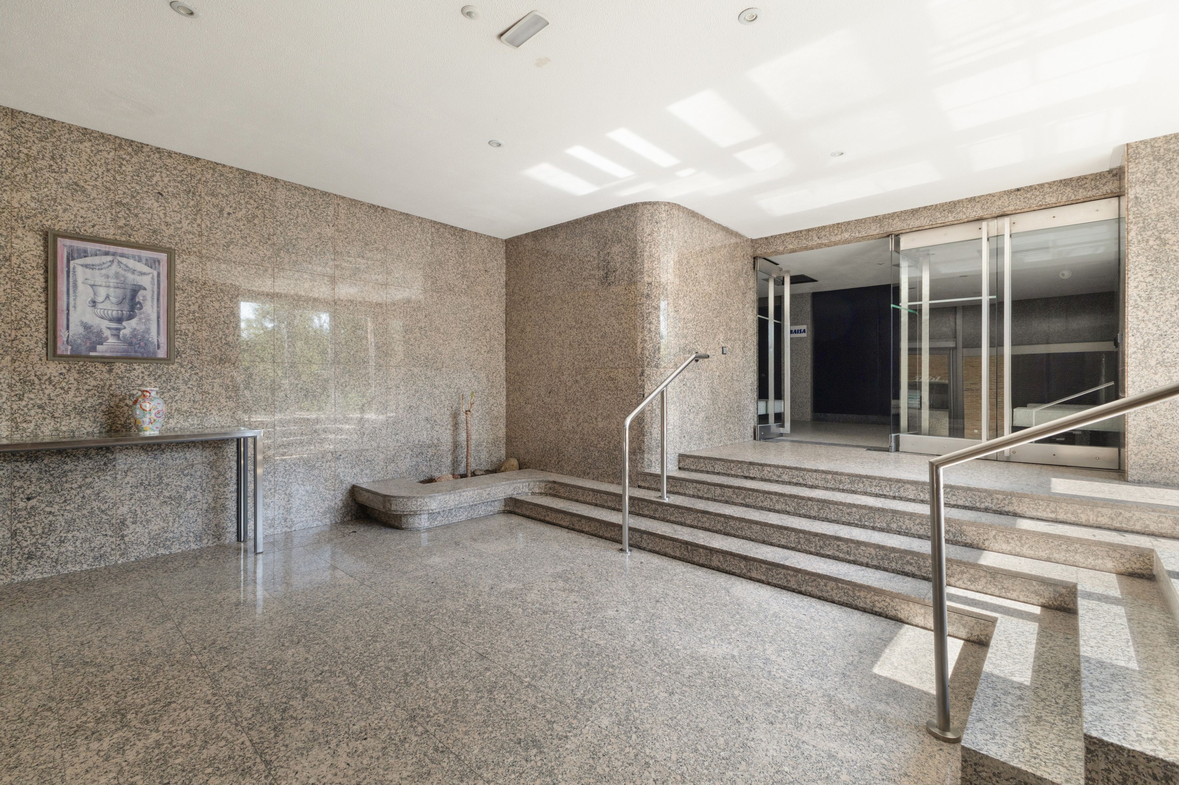 Un vestíbulo con piso de mármol, una escalera con barandilla de metal y una obra de arte enmarcada en la pared.