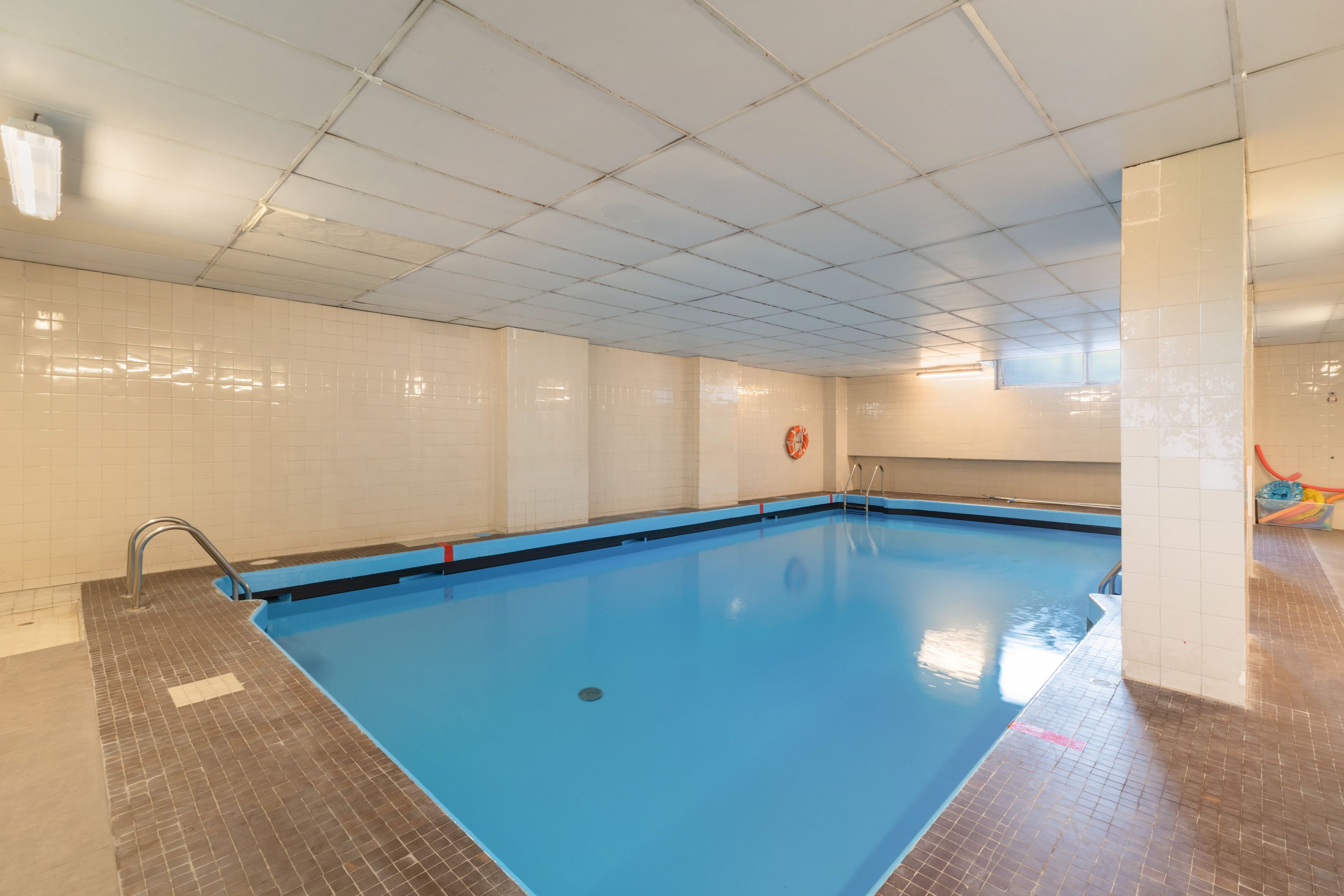 Una piscina interior con suelo y paredes de azulejos, un flotador en la pared y una escalera metálica que da acceso a la piscina.