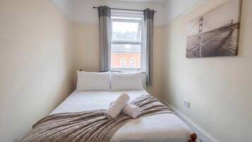 A bedroom with a bed, a window with curtains, a painting on the wall, and a view of a building outside.