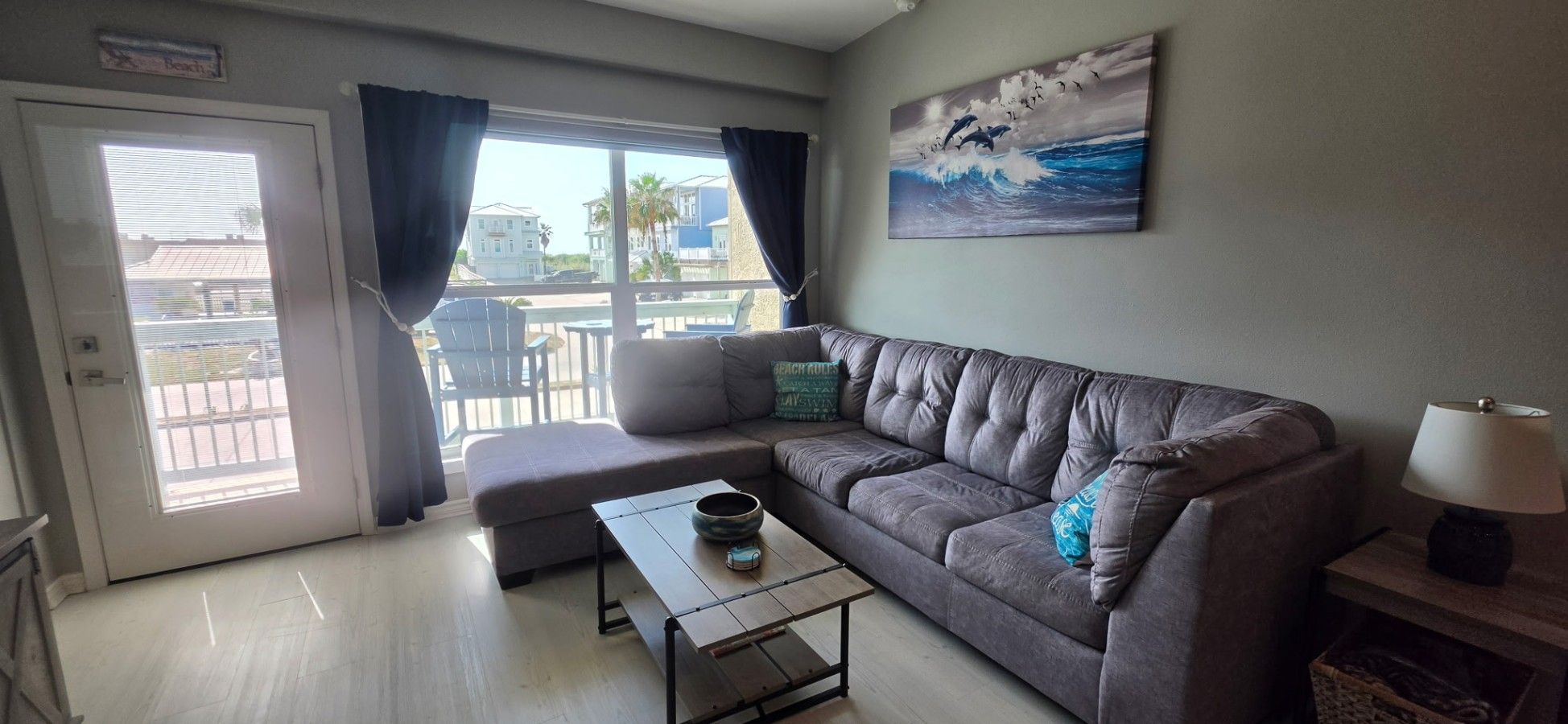 A living room with a grey sofa, a small table, a lamp, and a painting of dolphins on the wall.