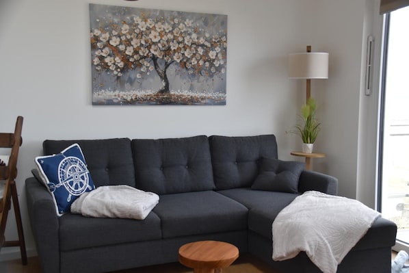 A living room with a dark grey sofa, a round wooden coffee table, a blue pillow with a white design, a white throw blanket, and a painting of a tree with white flowers on the wall.