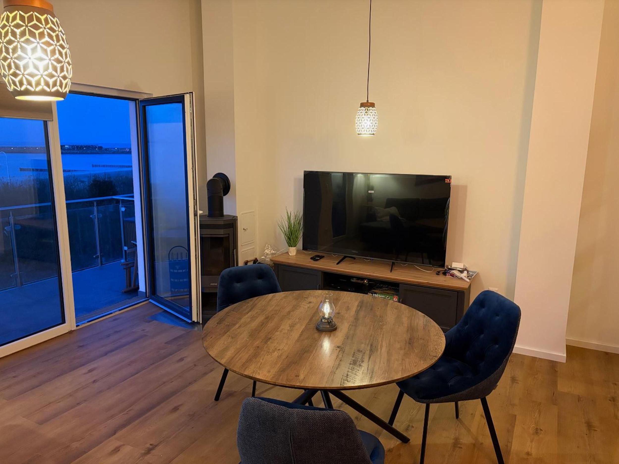 Ein modernes Wohnzimmer mit einem runden Holztisch, zwei blauen Stühlen, einem Flachbildfernseher und einer Glastür, die auf einen Balkon führt.