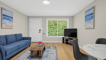 A living room with a blue sofa, a coffee table with a vase, a TV stand, a dining table, and a window with a view of greenery.