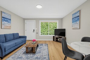 Una sala de estar con un sofá azul, una mesa de centro con un jarrón, un mueble para la televisión, una mesa de comedor y una ventana con vistas a zonas verdes.