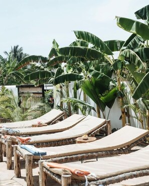 Una hilera de sillones de madera con cojines color beige, ubicada en un espacio al aire libre iluminado por el sol, con grandes plantas de banano y otra vegetación tropical en el fondo.