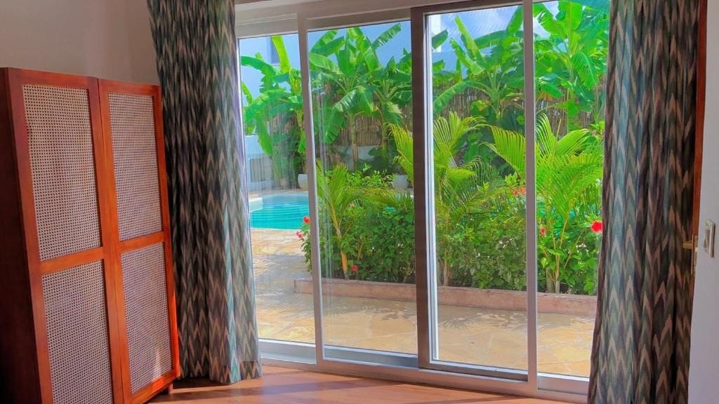 Una habitación con puerta corrediza de vidrio que da a un área de piscina con abundante vegetación.
