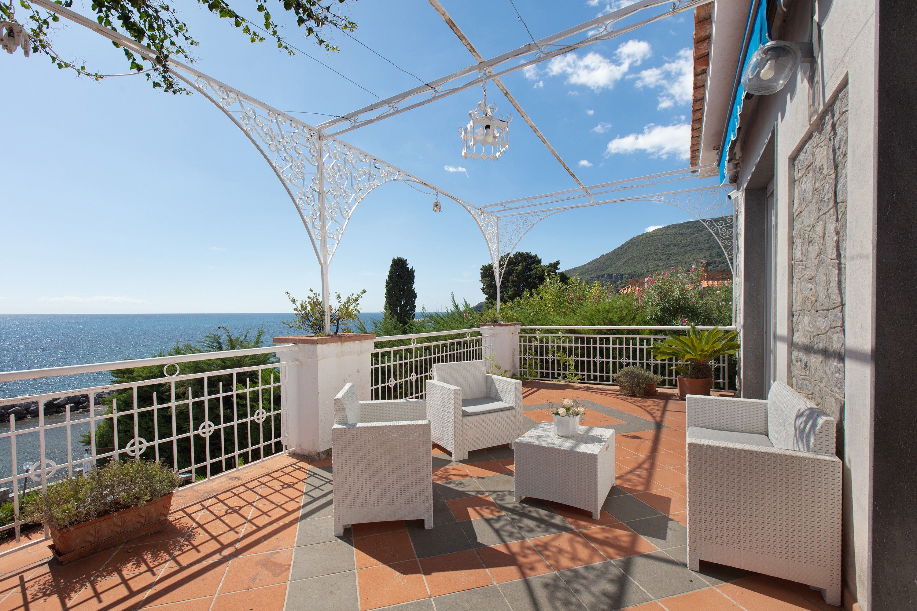 Una terraza con muebles de mimbre blanco, vista al mar y una montaña al fondo.