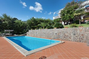 A swimming pool with a stone wall and tiled deck, surrounded by greenery and a building with a balcony. - Villa Scogliera dei Gabbiani with sea view, shared pool and Wi-Fi (Scario)