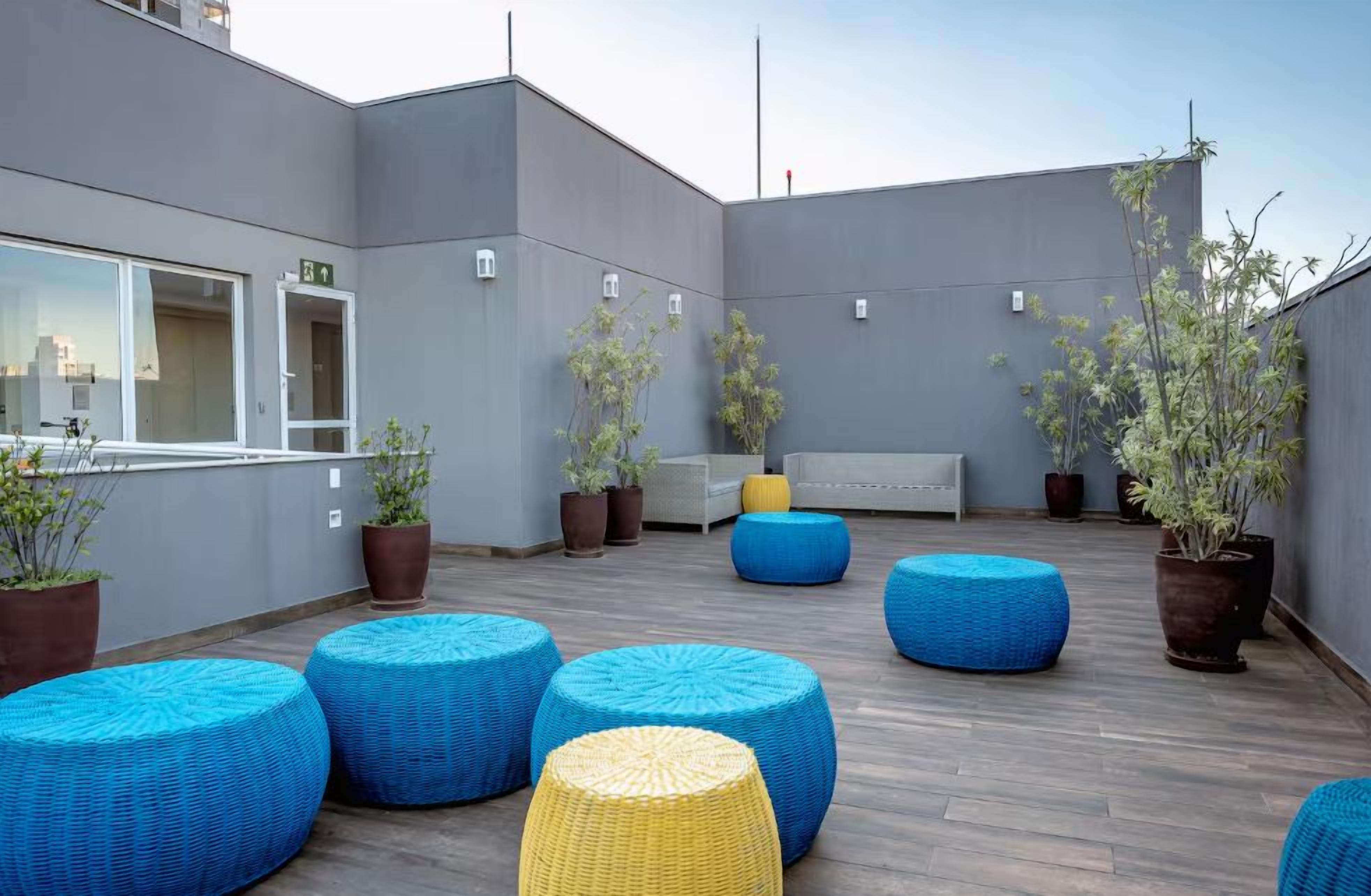 A rooftop terrace with blue and yellow woven seating, potted plants, and a white bench.