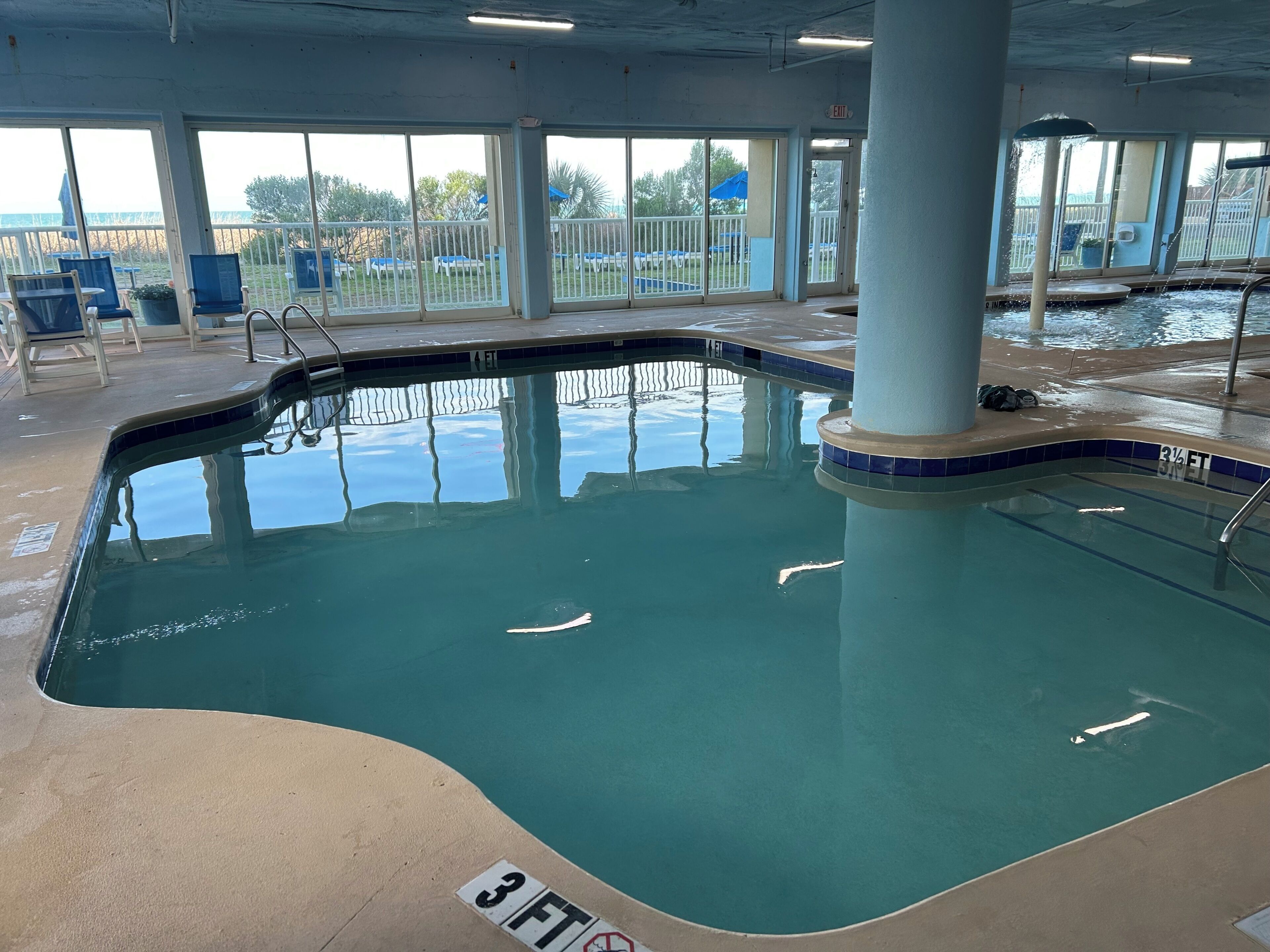 An indoor swimming pool with clear water, a 3ft depth marking, and a view of the outside through large windows.