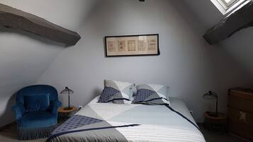 Une chambre avec un plafond en pente, un lit avec une literie à motifs, un fauteuil bleu et un tableau encadré au mur.