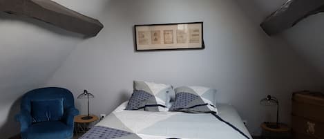 Ein Schlafzimmer mit schrägverdeckter Decke, einem Bett mit gemusterten Textilien, einem blauen Sessel und einem gerahmten Bild an der Wand.