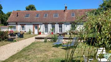 Een huis met twee verdiepingen, een pannendak, een grindpad en een tuin met potplanten en tuinmeubilair.