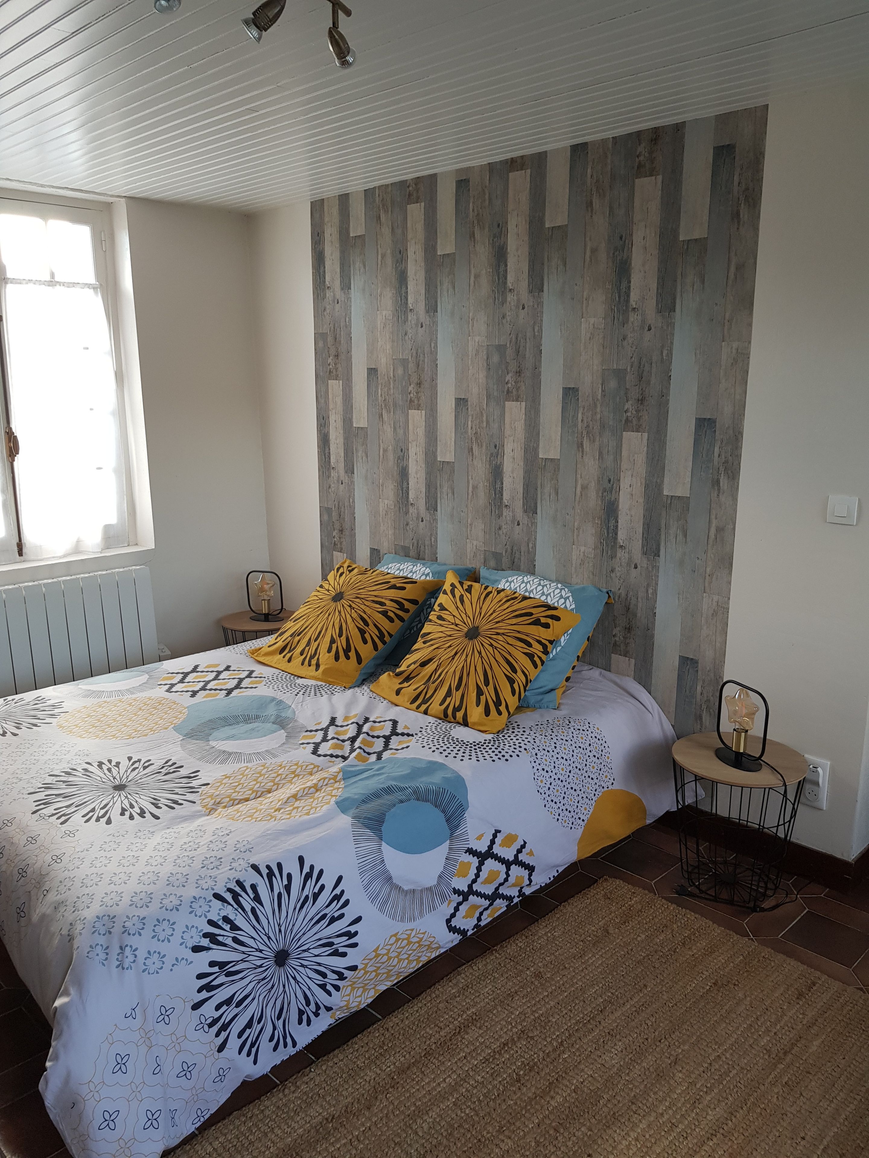 Een slaapkamer met een bed, een raam, een wand met houten panelen en een kleine tafel met een lamp.