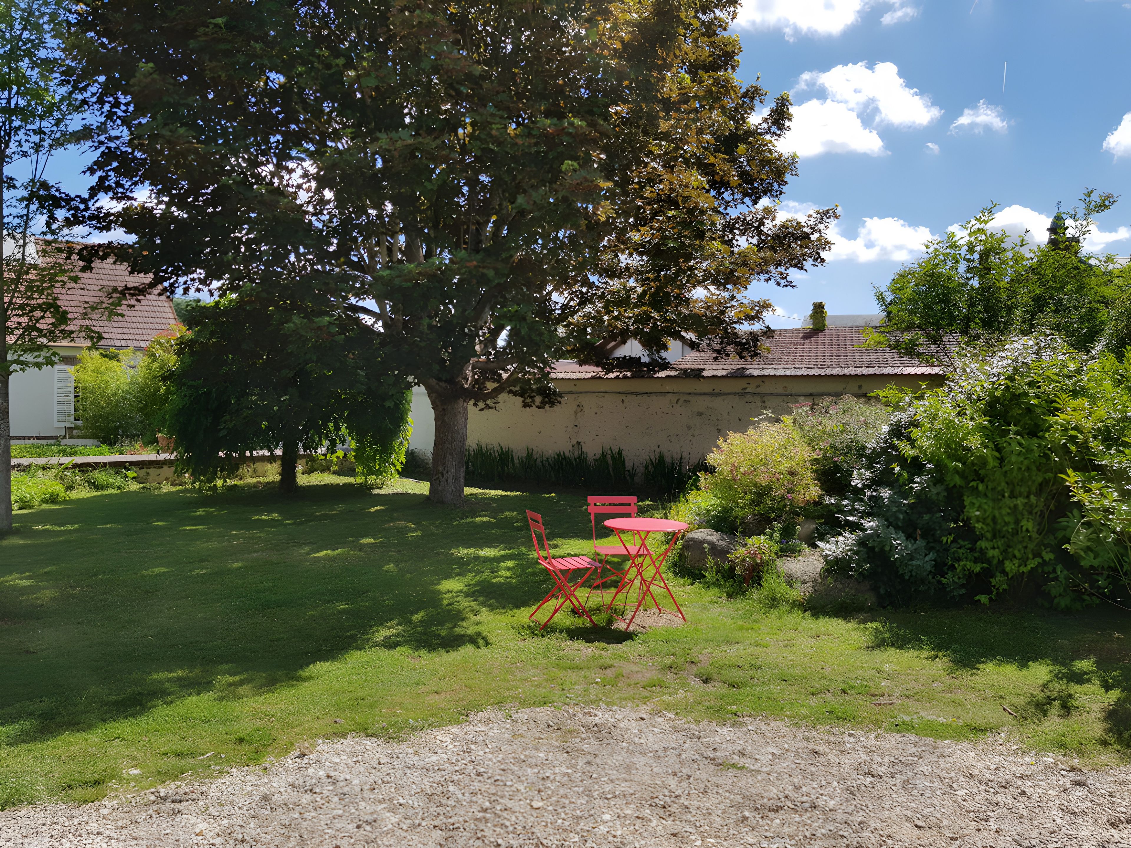 Un jardin avec un arbre, un chemin de gravier et une table et des chaises rouges.