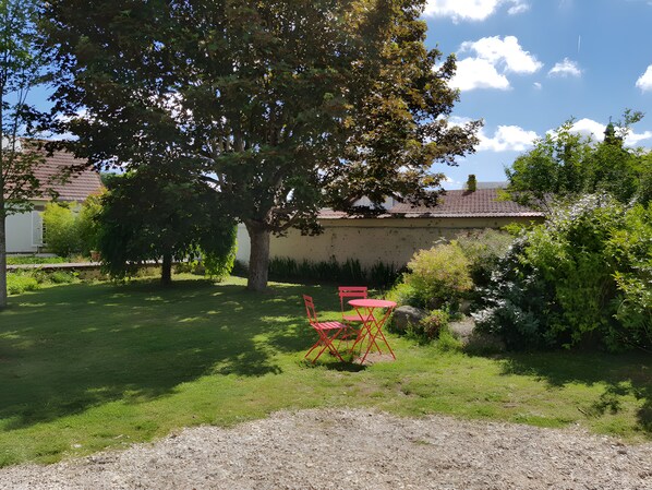 A garden with a tree, a gravel path, and a red table and chairs.