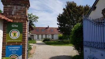 Une entrée fermée par un portail mène à un bâtiment à la façade en briques. Un chemin de gravier conduit à l’entrée, et des panneaux indiquent « Gîte » et « CHAMBRES D’HÔTES ».