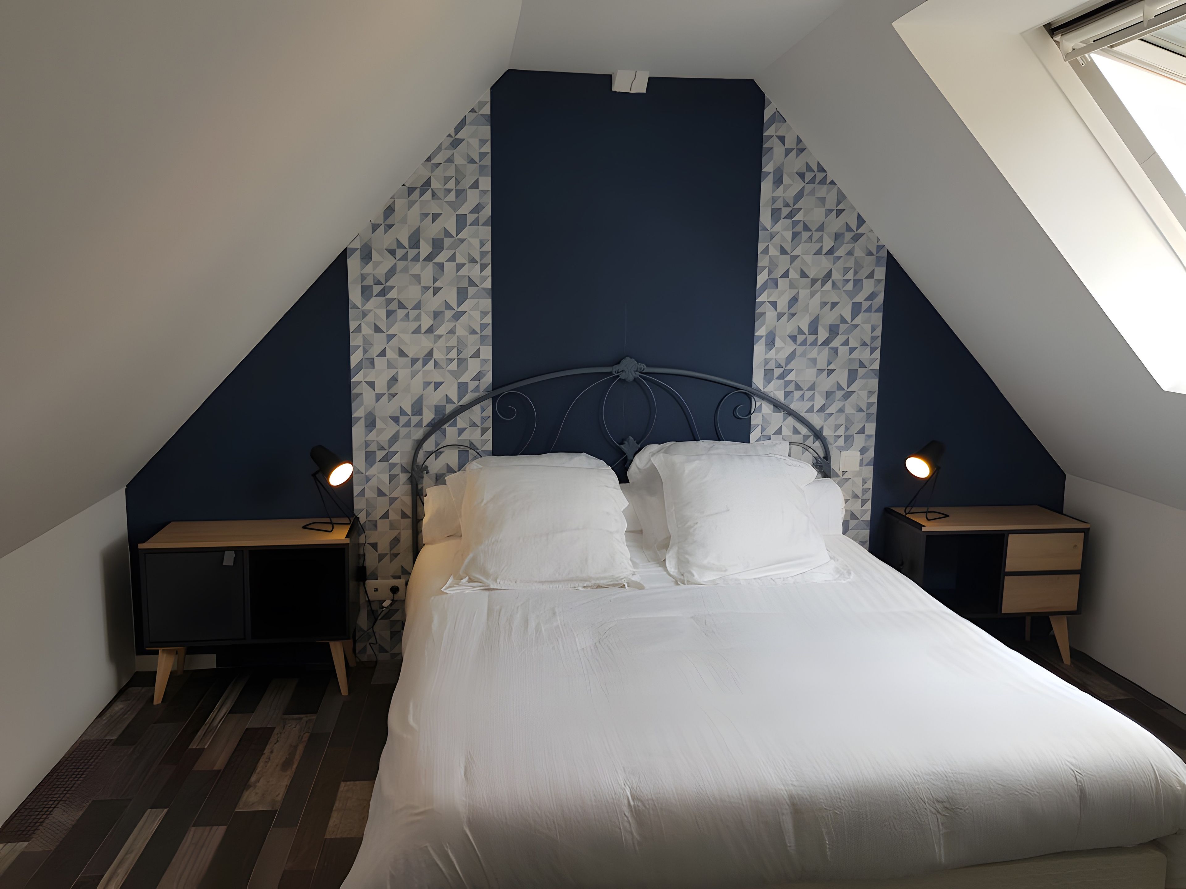 Un dormitorio con techo inclinado, una cama con ropa blanca, dos mesitas de noche con lámparas y paredes con diseño geométrico.