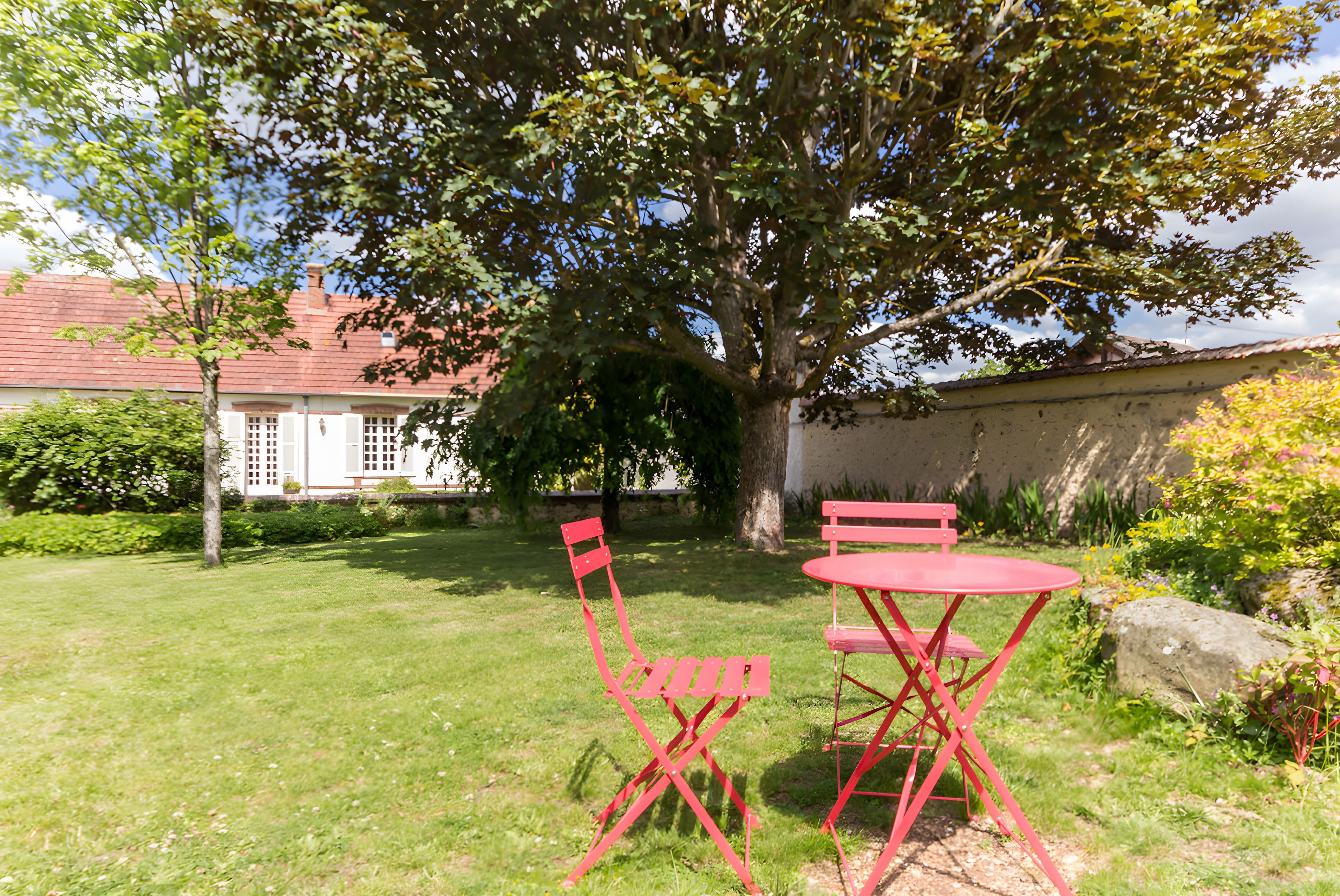 Een tuin met een rode metalen tafel en stoelen, een boom en een gebouw met een rood dak op de achtergrond.
