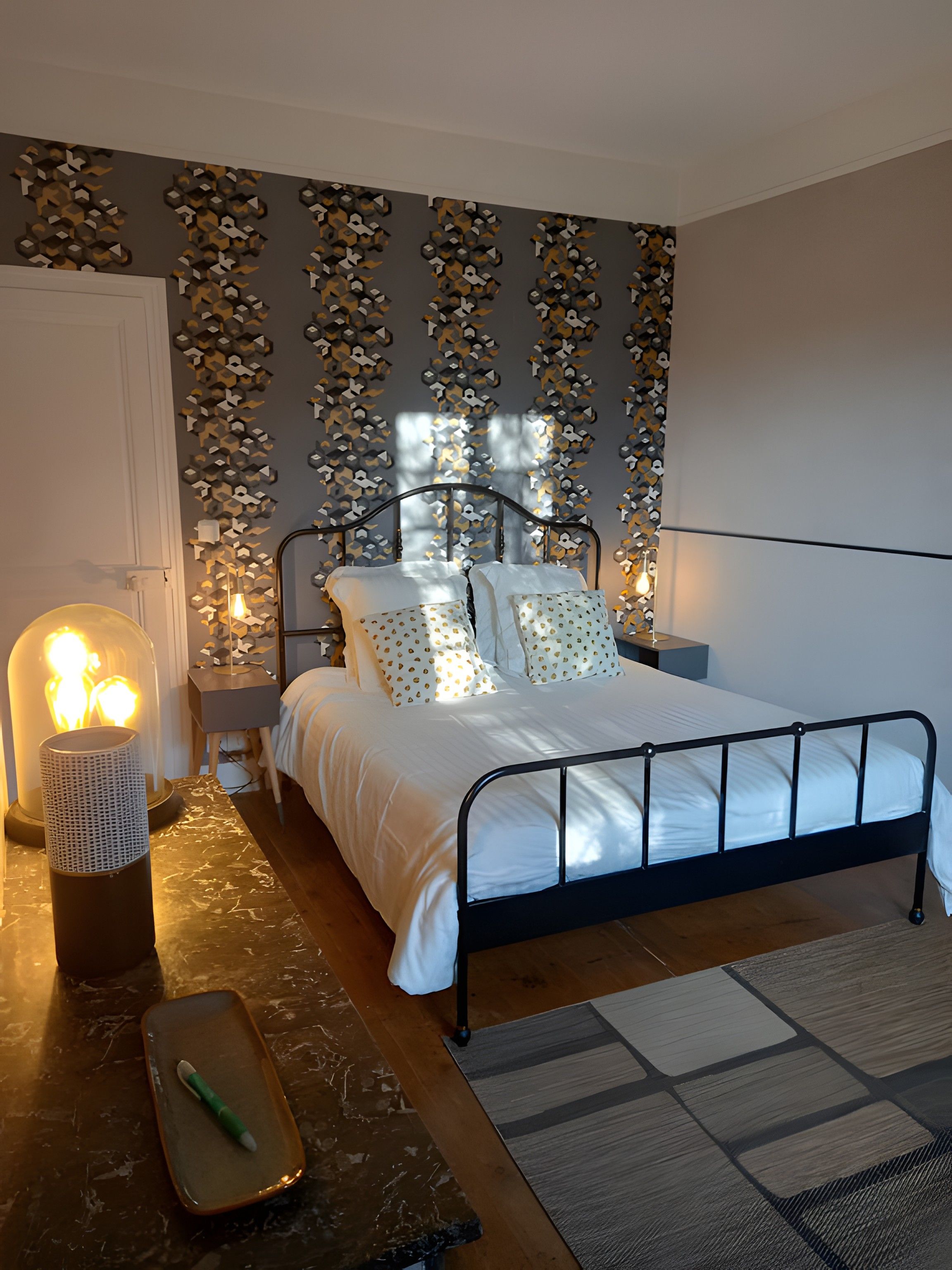 Un dormitorio con estructura de cama metálica, ropa de cama blanca, pared estampada y una lámpara de noche.