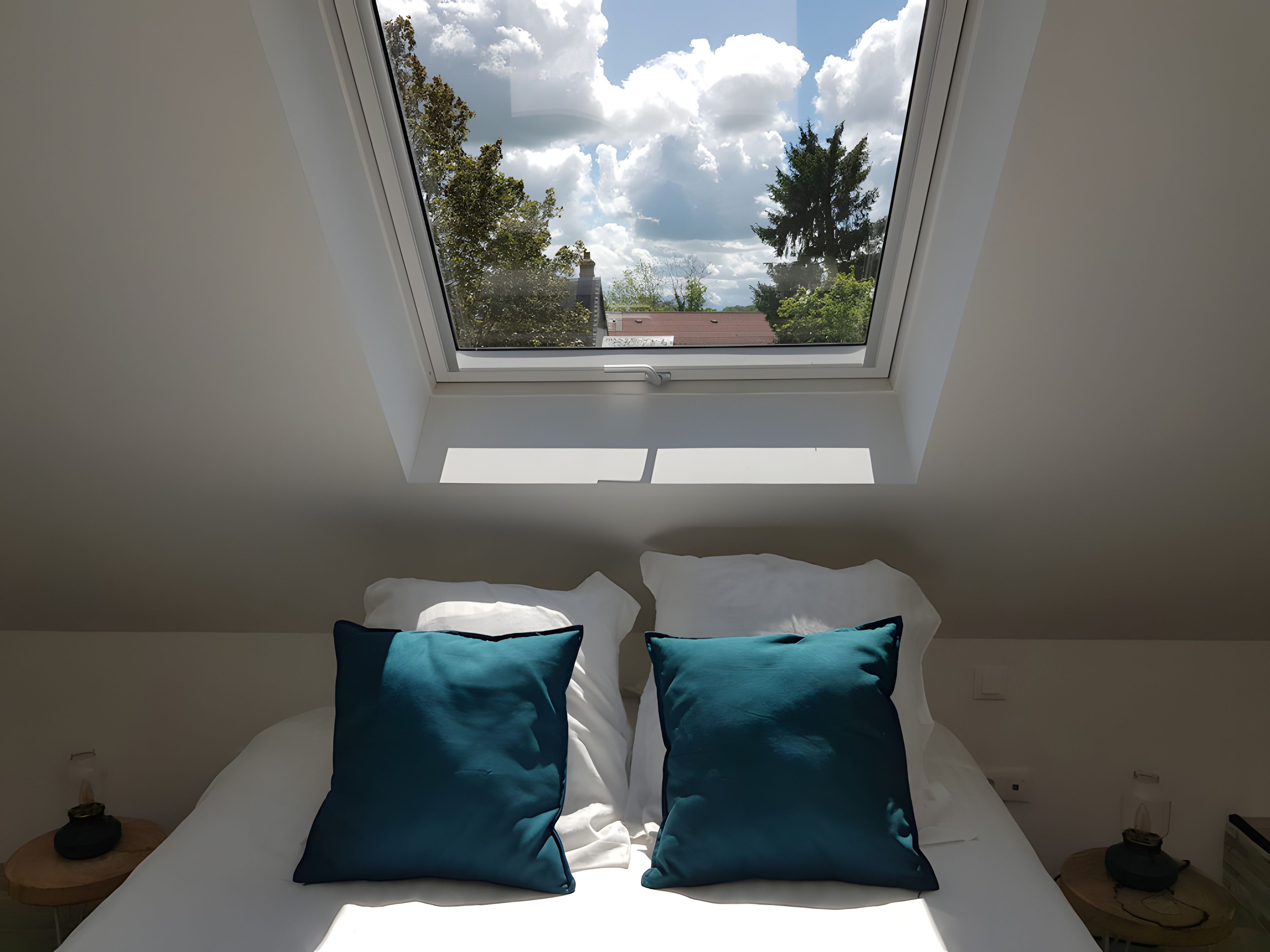 Een kamer met dakraam, een bed met witte en teal kussens, en uitzicht op bomen en een gebouw.