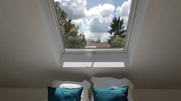 Een kamer met dakraam, een bed met witte en teal kussens, en uitzicht op bomen en een gebouw.