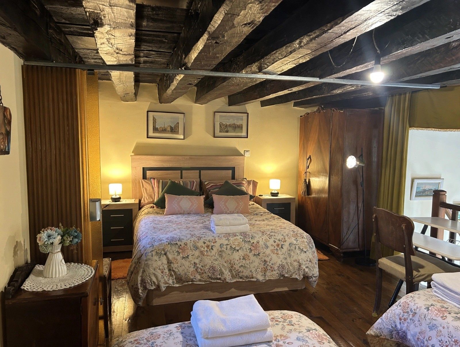 Une chambre avec un plafond en bois, un lit recouvert d’une literie à motifs floraux, des lampes de chevet et une armoire en bois.