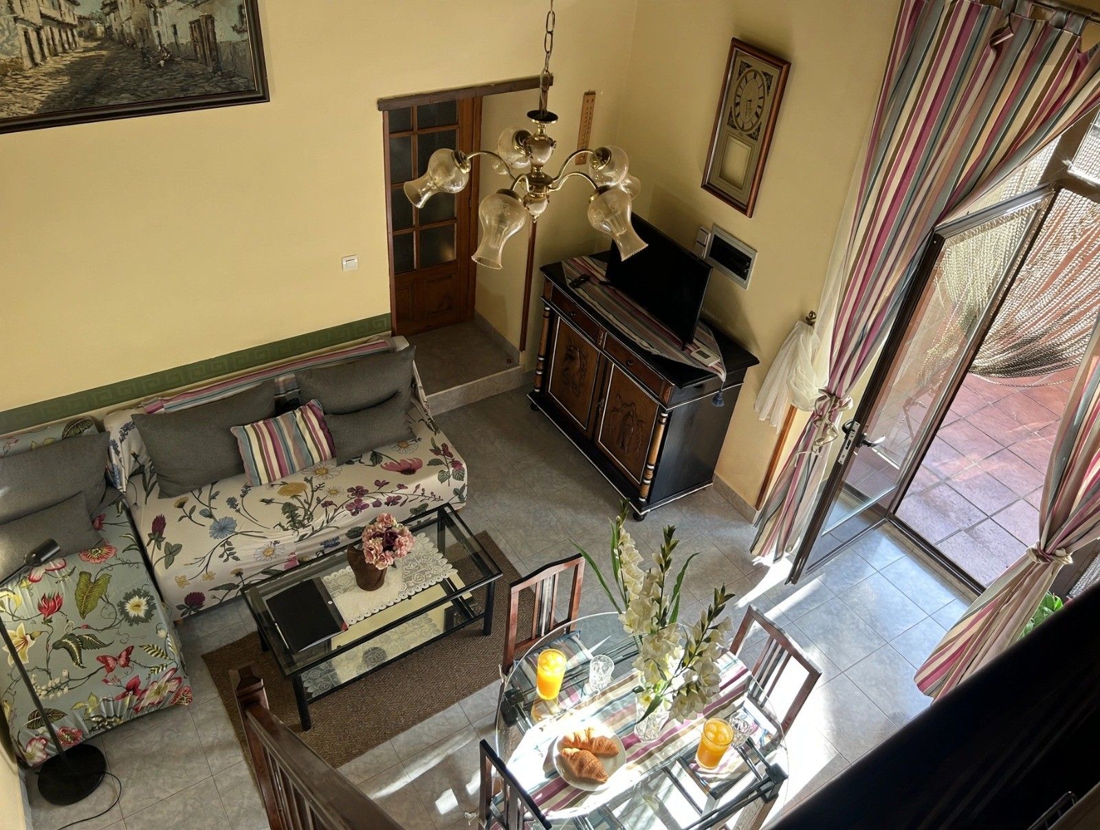 Un soggiorno con divano, tavolino in vetro e tavolo da pranzo con sedie. C'è una televisione su un mobile, un lampadario e una finestra con tende.